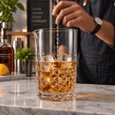 A clear cocktail mixing pitcher filled with amber liquor and large ice cubes, being stirred by a person on a marble bar counter. A bottle, shaker, and bar decor are visible in the background.