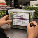Person holding a container of arugula with a label on a kitchen counter