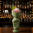 A green ceramic tiki-style drinkware piece on a wooden bar counter, filled with crushed ice and a pink drink, garnished with a lime wheel, mint, and a black straw, with a blurred bar shelf of bottles in the background.