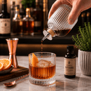 Person pouring bitters into a cocktail with a bottle of bitters and ingredients in the background.