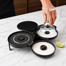 A black margarita glass rimmer organizer on a white marble countertop, with a hand lifting a glass in use. labeled salt, sugar sand lime juice.