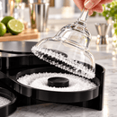 A hand holds a clear Margarita glass rimmed with white salt crystals above a black salt dish filled with coarse white salt, set on a marble countertop with limes and bar tools blurred in the background.