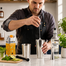A man in a dark shirt and apron stirs a cocktail shaker on a kitchen counter, with a bottle of premium rum, a carton of mojito, lime slices, mint, and metal bar tools nearby.
