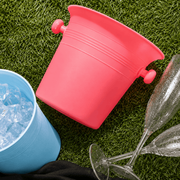 A bright pink metal ice bucket with side handles, shown on artificial grass next to a blue bucket filled with ice, two clear champagne flutes, and a black cloth.
