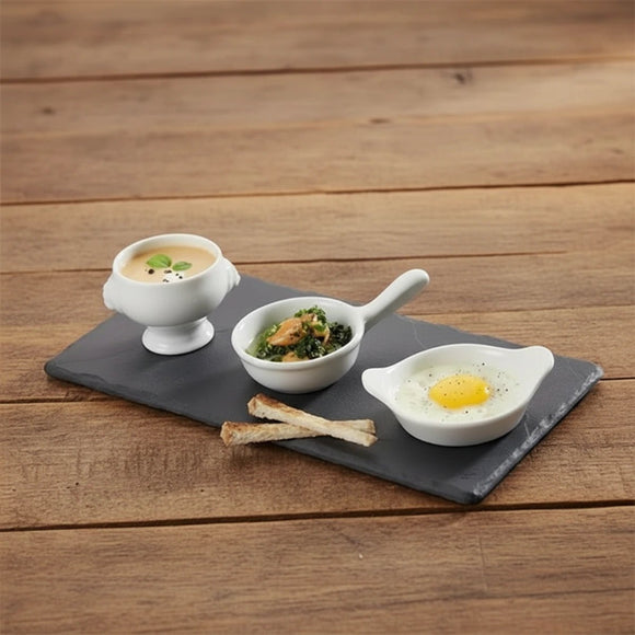 Three small white bowls with different dishes on a dark slate tray, placed on a wooden surface.
