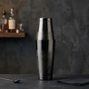A Gunmetal cocktail shaker with a matching lid, standing upright on a dark tabletop against a textured charcoal background. A wooden shelf with bottles is blurred in the background, along with a spoon and a folded dark cloth.