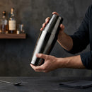 A BarBits Gunmetal cocktail shaker being held by a bartender's hands in a dimly lit bar setting with dark gray walls and wooden shelves in the background.