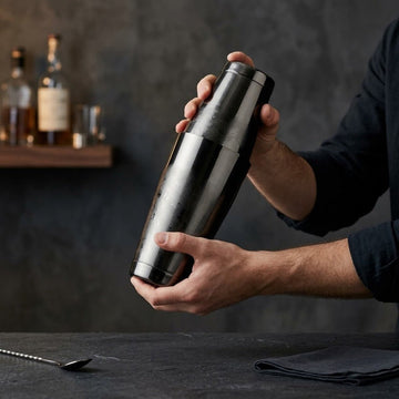 A BarBits Gunmetal cocktail shaker being held by a bartender's hands in a dimly lit bar setting with dark gray walls and wooden shelves in the background.