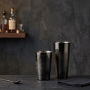 Two Gunmetal metallic cocktail shakers on a dark tabletop against a textured charcoal wall, with a blurred shelf of bottles in the background.