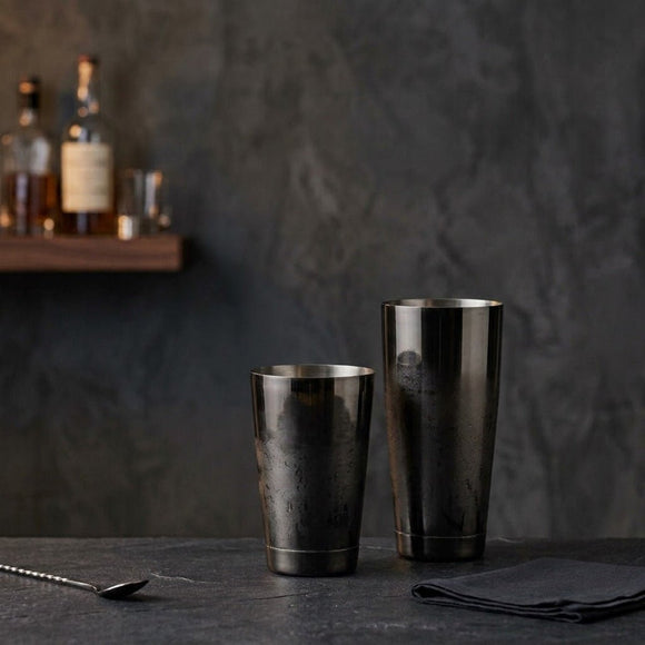 Two Gunmetal metallic cocktail shakers on a dark tabletop against a textured charcoal wall, with a blurred shelf of bottles in the background.