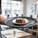 Person holding a black tray with a dessert plate featuring a lava cake, ice cream, and berries in a restaurant setting.