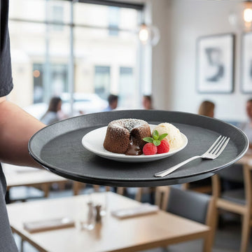 Person holding a black tray with a dessert plate featuring a lava cake, ice cream, and berries in a restaurant setting.