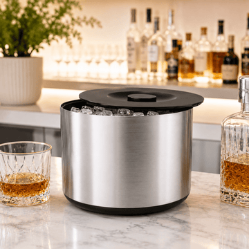 Metallic ice bucket on a marble surface with glasses of whiskey and bottles in the background.