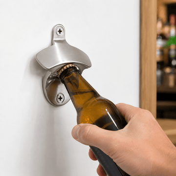 A hand using a wall-mounted silver bottle opener to open an amber glass beer bottle against a white wall.