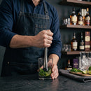 A bartender in a dark denim apron mudding lime and mint garnish into a clear rocks glass on a dark bar counter, with shelves of liquor bottles in the background.