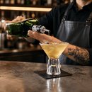 A bartender in a black apron pours a clear liquid from a green bottle into a martini glass filled with a golden cocktail and ice, set on a dark bar counter.