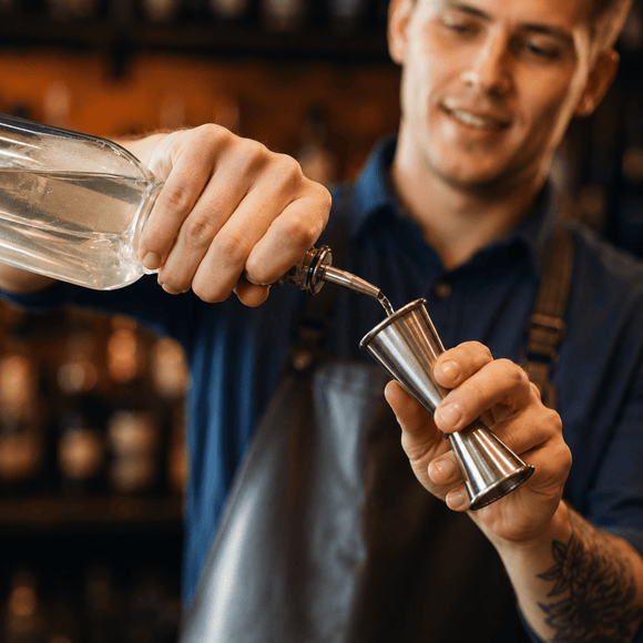 A bartender in a navy blue shirt and dark apron pours a clear liquid from a bottle into a stainless steel jigger, with a blurred bar background.