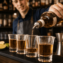 A bartender in a dark blue shirt and gray apron pours a clear amber liquid from a bottle into four clear shot glasses arranged on a black bar mat, with limes and blurred bar shelves in the background.