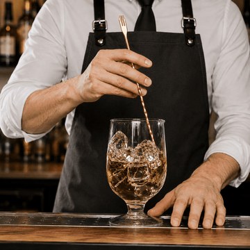 A bartender in a black apron and white shirt stirs a glass of amber-colored cocktail with ice cubes using a copper-toned bar spoon on a wooden bar counter.
