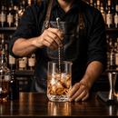 A bartender in a black shirt and dark leather apron stirs a glass of amber liquor with ice in a bar setting, with bottles and bar tools visible in the background.