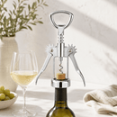 A polished stainless steel wine bottle opener with a corkscrew is shown positioned over the neck of a dark wine bottle, with a glass of white wine and green grapes in the softly lit background.