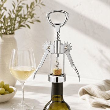 A polished stainless steel wine bottle opener with a corkscrew is shown positioned over the neck of a dark wine bottle, with a glass of white wine and green grapes in the softly lit background.