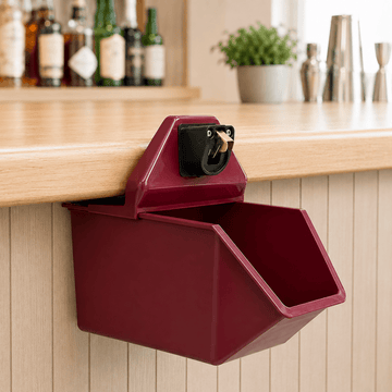A dark red wall-mounted dispenser with a black dial and gold handle, mounted on a light wood bar counter in a bar setting.