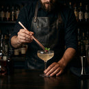 A bartender in a black leather apron garnishes a cocktail in a coupe glass with a copper-colored metal stirrer, set against a dark, blurred bar background.