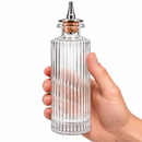 A hand holding a Victorian Glass Bitters Bottle 145ml with a cork stopper and metal pour spout, shown against a plain white background.