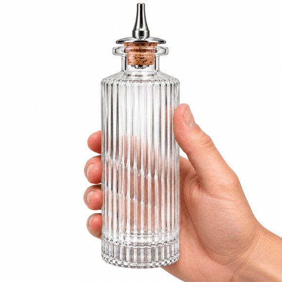 A hand holding a Victorian Glass Bitters Bottle 145ml with a cork stopper and metal pour spout, shown against a plain white background.