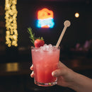 Hand holding a pink cocktail with ice, a strawberry, and a rosemary sprig against a dark background with colorful lights.