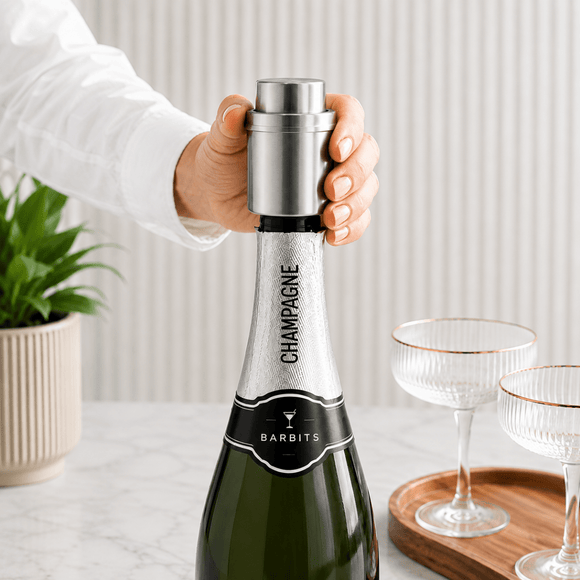 A hand in a white shirt holds a silver metal champagne stopper onto a dark green champagne bottle labeled 'CHAMPAGNE' and 'BARBITS'. Two clear coupe glasses with gold trim sit on a wooden tray in the background on a light marble countertop.