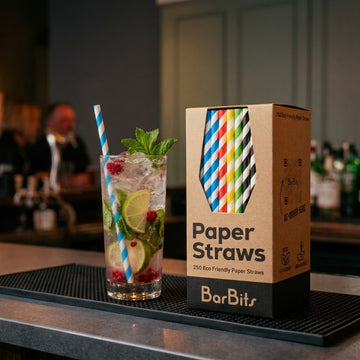 A BarBits paper straws product box stands on a bar counter next to a tall cocktail glass with a blue-and-white striped paper straw. The box shows a colorful tie-shaped window with striped paper straw illustrations and the text 'Paper Straws' and 'BarBits'.