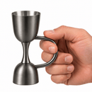 A hand holding a small double- jigger cocktail measure brushed dark brushed finish against a plain white background.
