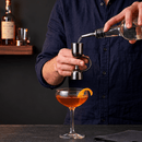 A bartender in a navy blue shirt uses a stainless steel jigger to measure liquid from a clear bottle into a coupe glass containing an orange cocktail, with whiskey bottles on a shelf in the background.