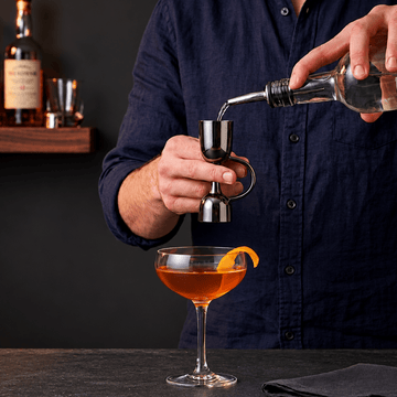 A bartender in a navy blue shirt uses a stainless steel jigger to measure liquid from a clear bottle into a coupe glass containing an orange cocktail, with whiskey bottles on a shelf in the background.