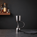 A polished stainless steel cocktail mixing cup with a curved handle and integrated base, standing on a dark countertop against a dark gray wall. A wooden shelf with bottles is blurred in the background, and a folded dark cloth sits to the right.